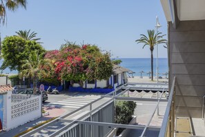 A balcony with a view of the beach, palm trees, and a building with a blue and white facade.