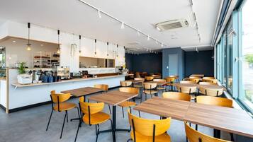 A modern cafe with wooden tables and yellow chairs, a counter with kitchen appliances, and large windows offering a view.