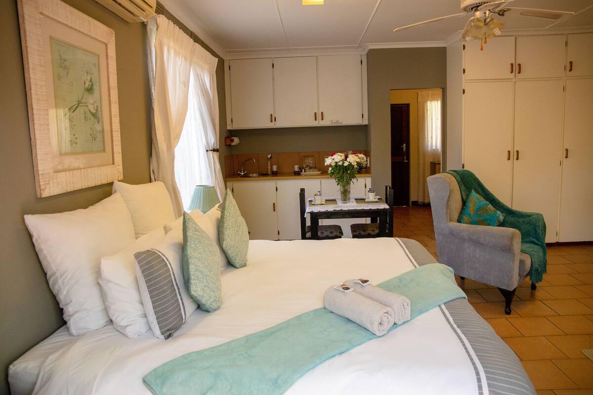 A hotel room with a bed, a chair, a table with a vase of flowers, and a kitchenette in the background.
