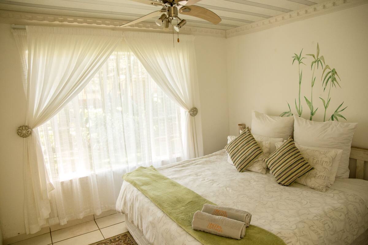 A bedroom with a bed, a ceiling fan, sheer curtains, a green throw blanket, and a potted plant.