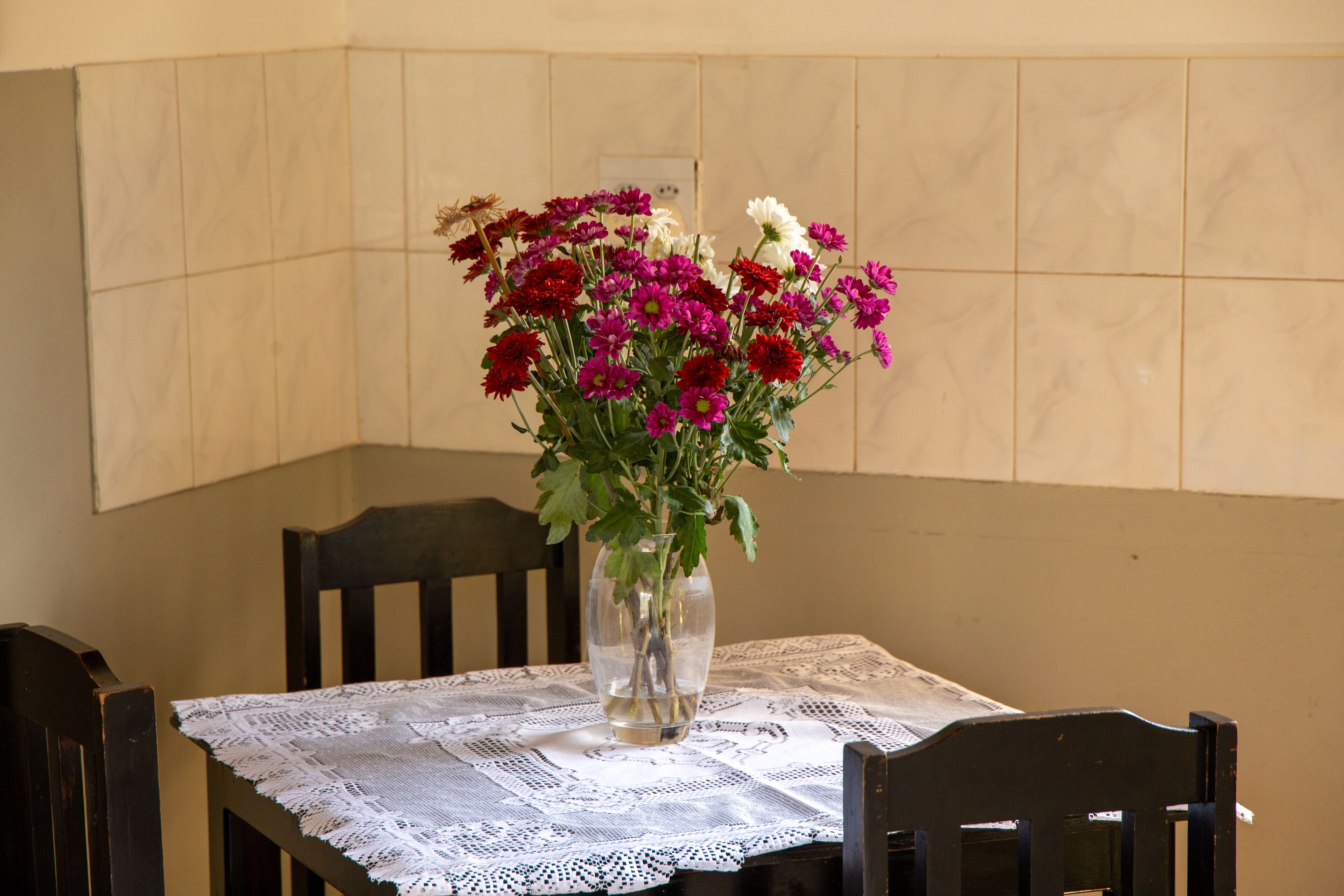 A dining table with a lace tablecloth, a vase with flowers, and wooden chairs.