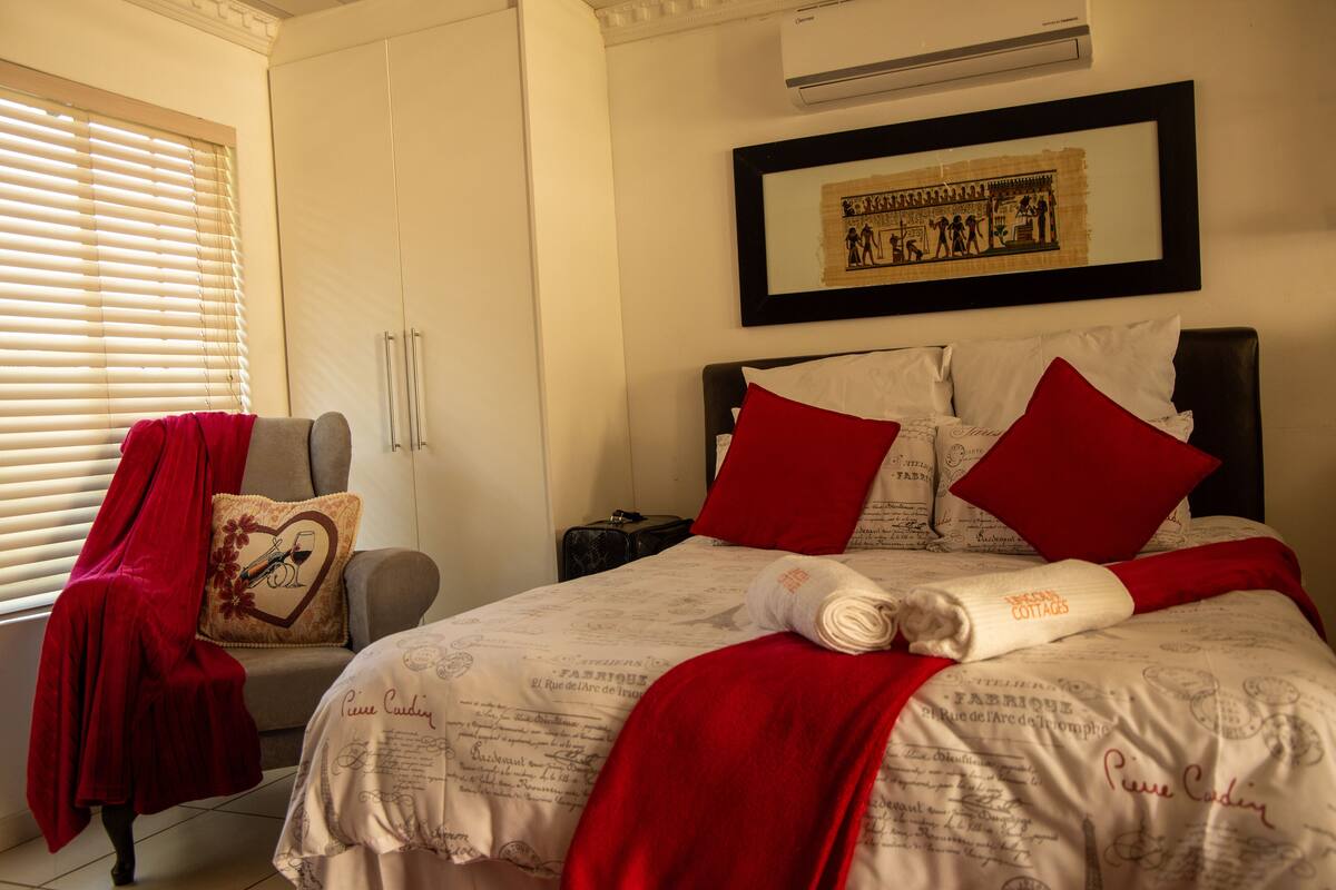 A neatly made bed with red pillows and a rolled towel, a chair with a red blanket, and a framed picture on the wall.