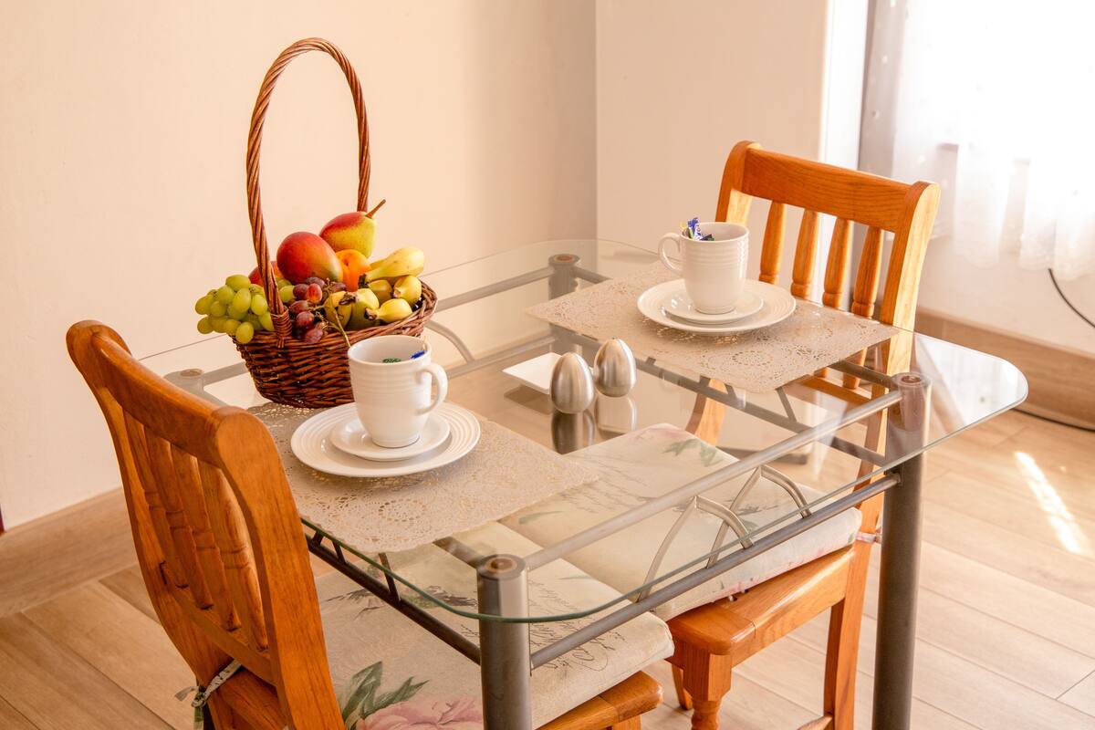 A glass-top dining table with two wooden chairs, a wicker fruit basket, white cups, saucers, and a small bowl.