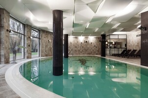 An indoor swimming pool with a clear view of the surrounding area, including a seating area with lounge chairs and a view of the outdoors through large windows.