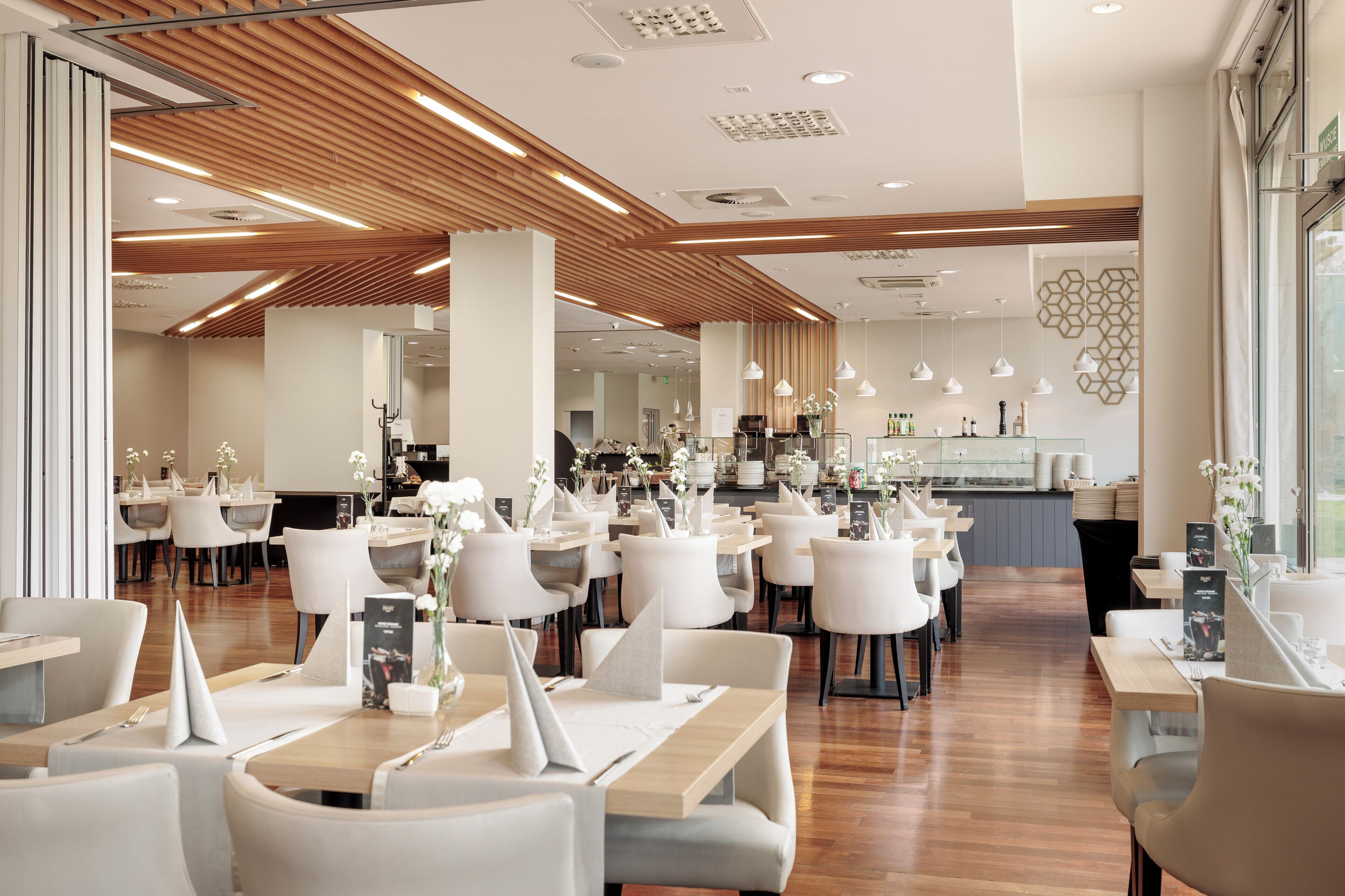 Een modern restaurant met witte stoelen en tafels, houten plafondpanelen en hanglampen.