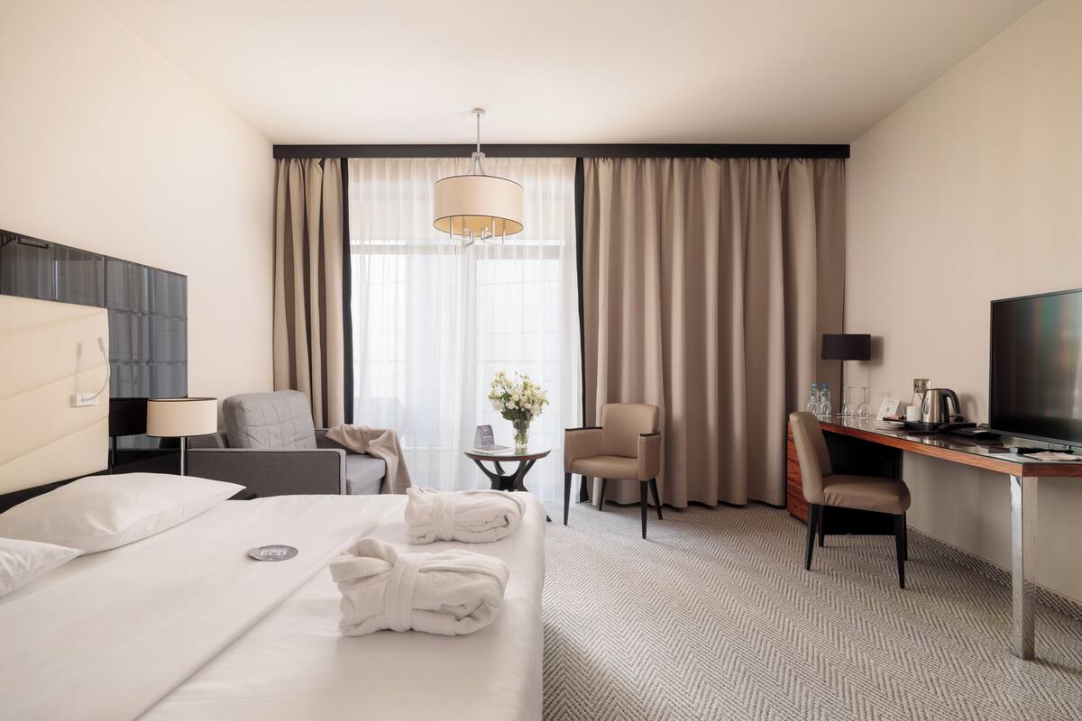 A hotel room with a bed, two chairs, a desk, a television, and a window with curtains.