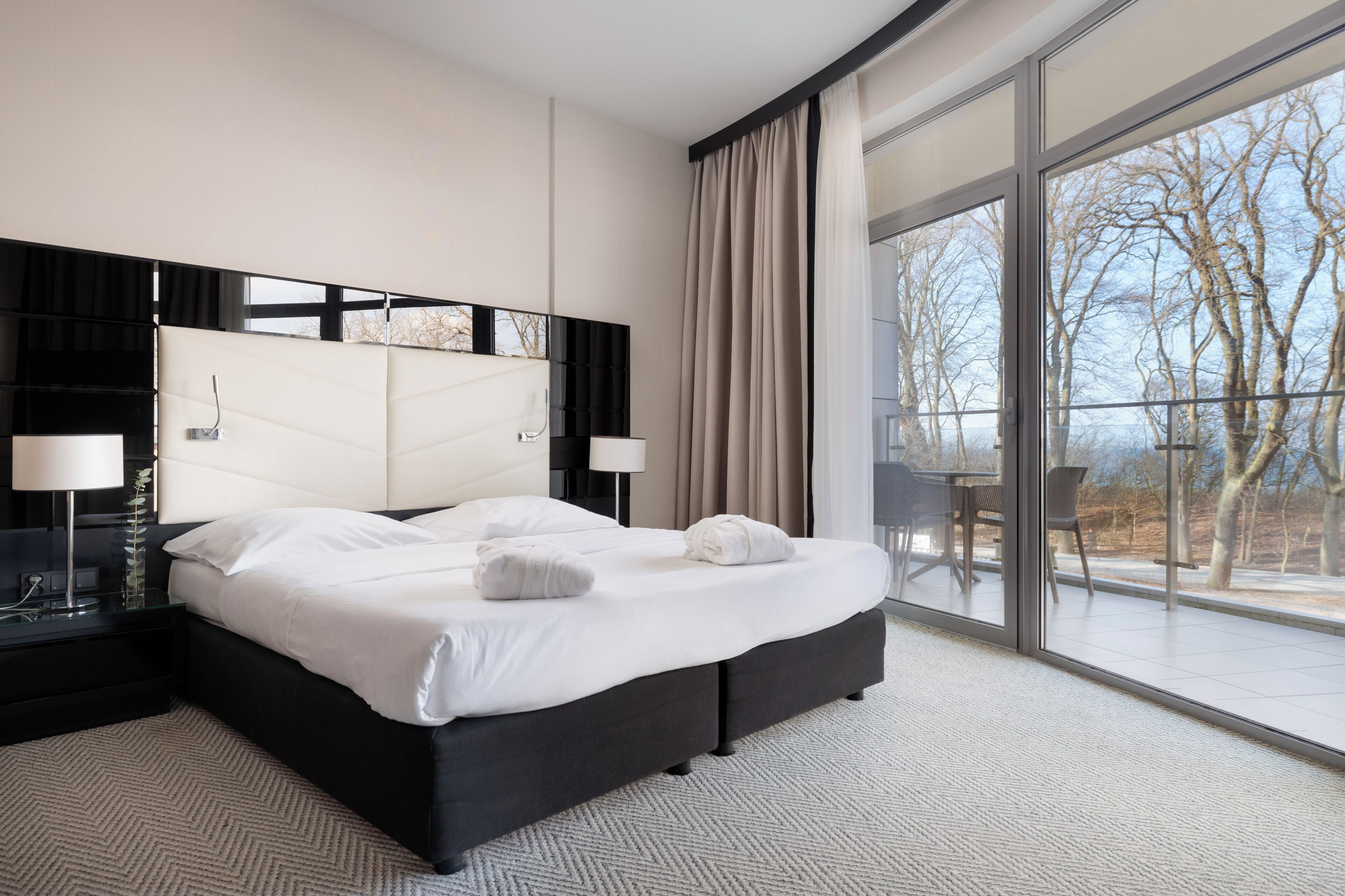 Ein modernes Hotelzimmer mit einem großen Bett, zwei Nachttischen, einem großen Fenster mit Blick auf Bäume und einem Balkon.