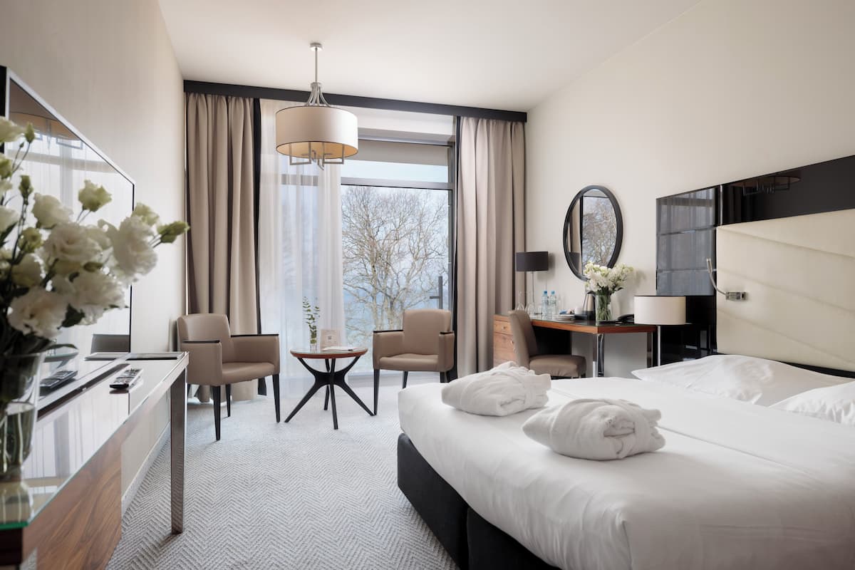 A hotel room with a bed, desk, chairs, a desk lamp, a mirror, and a window with curtains.
