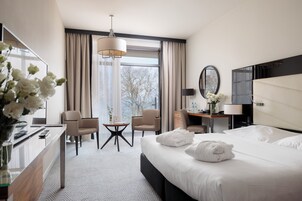 Quarto de hotel com cama, escrivaninha, cadeiras, abajur, espelho e janela com cortinas.