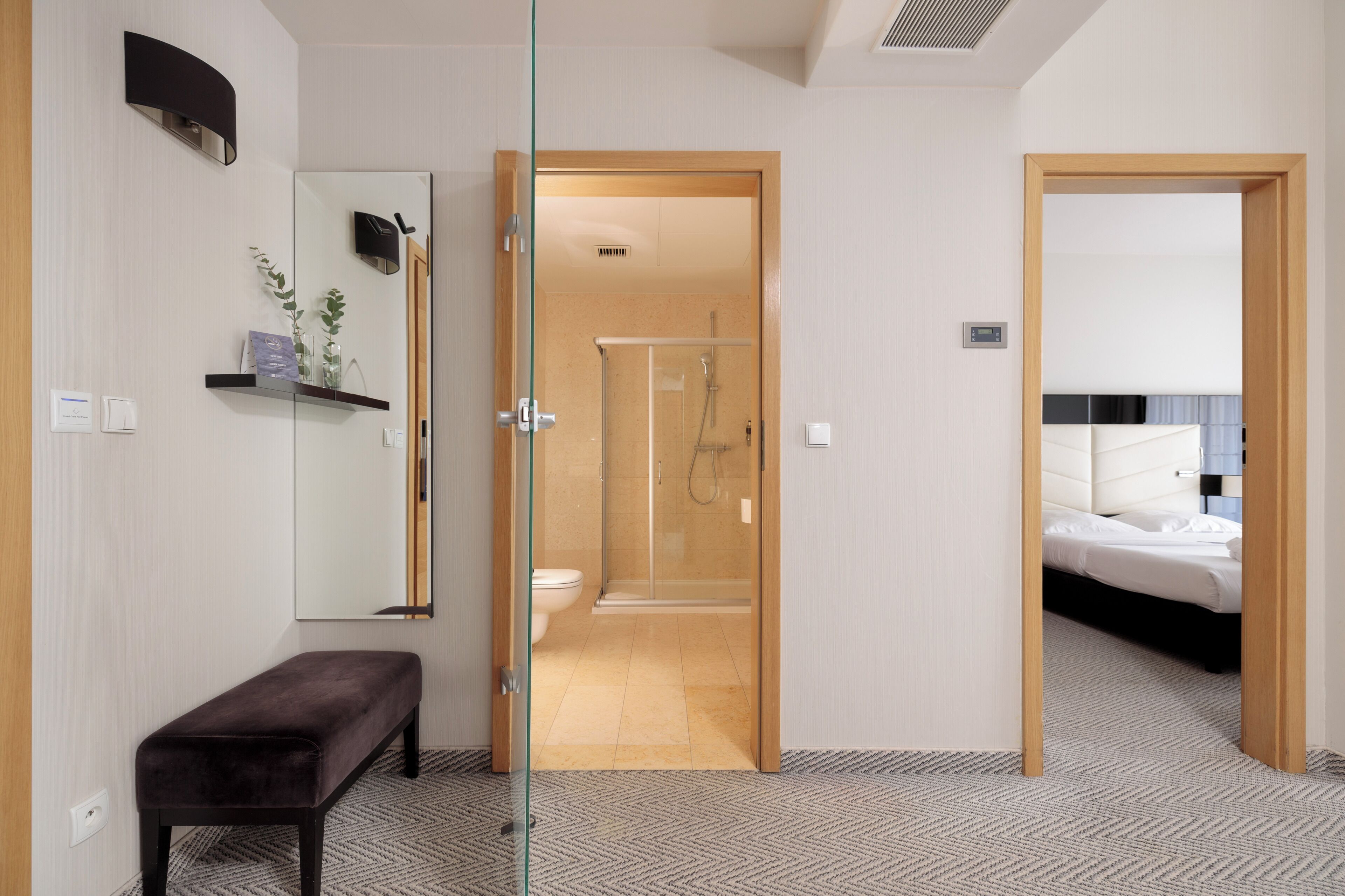 Een hotelkamer met een glazen deur naar een douchegedeelte, een nachtkastje en een bed met hoofdeinde.
