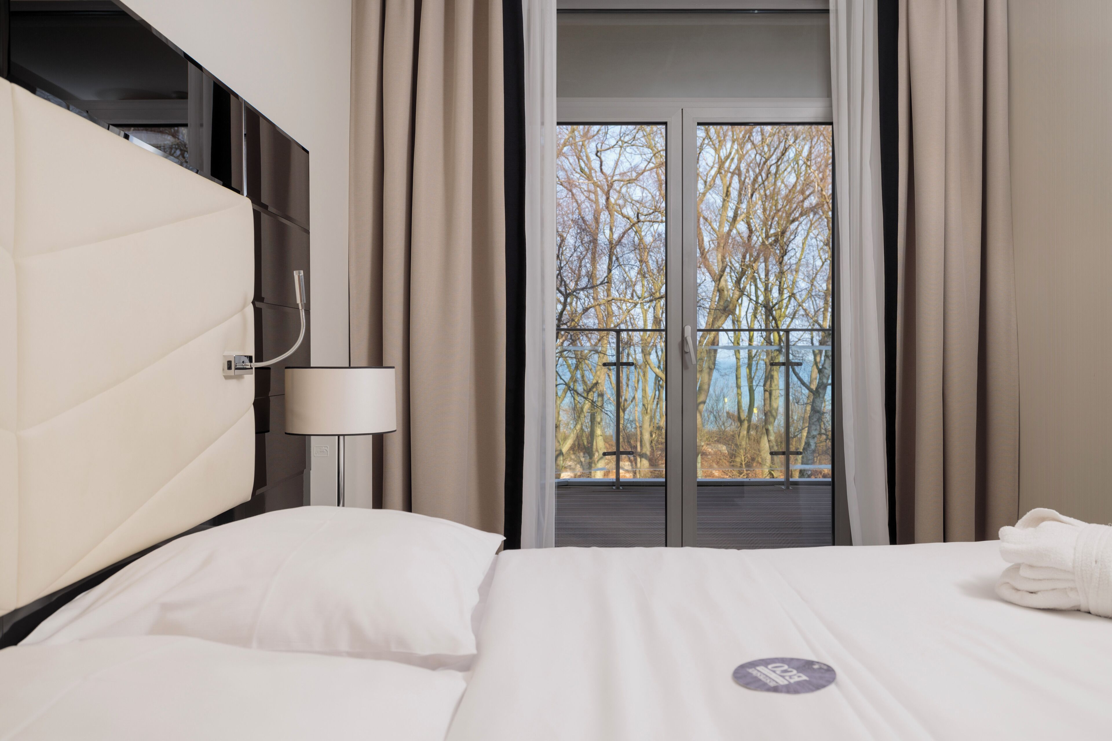 A hotel room with a bed, a wall-mounted lamp, a window with curtains, and a view of trees.