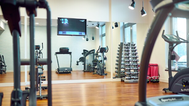A gym with various exercise equipment, a TV mounted on the wall, and a window with blinds. - Diune Hotel (Kolobrzeg)