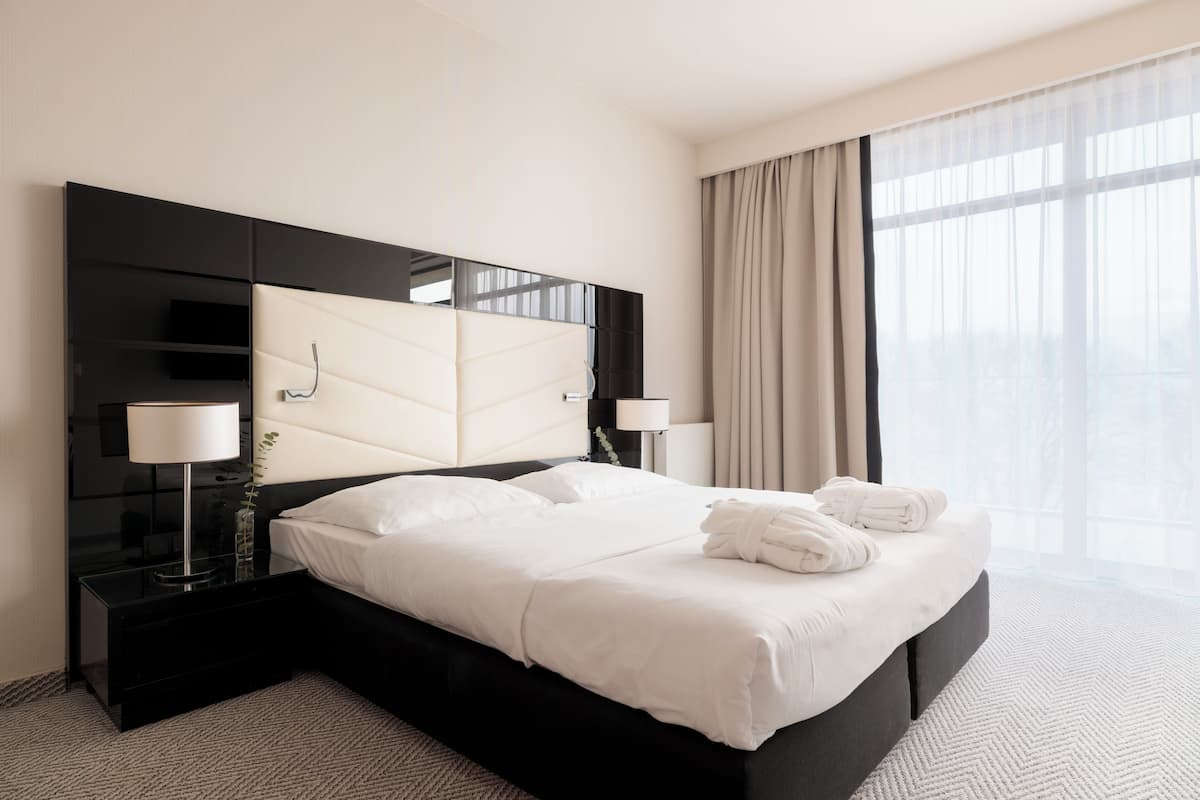 A modern hotel room with a large bed, two bedside tables, and a large window with curtains.