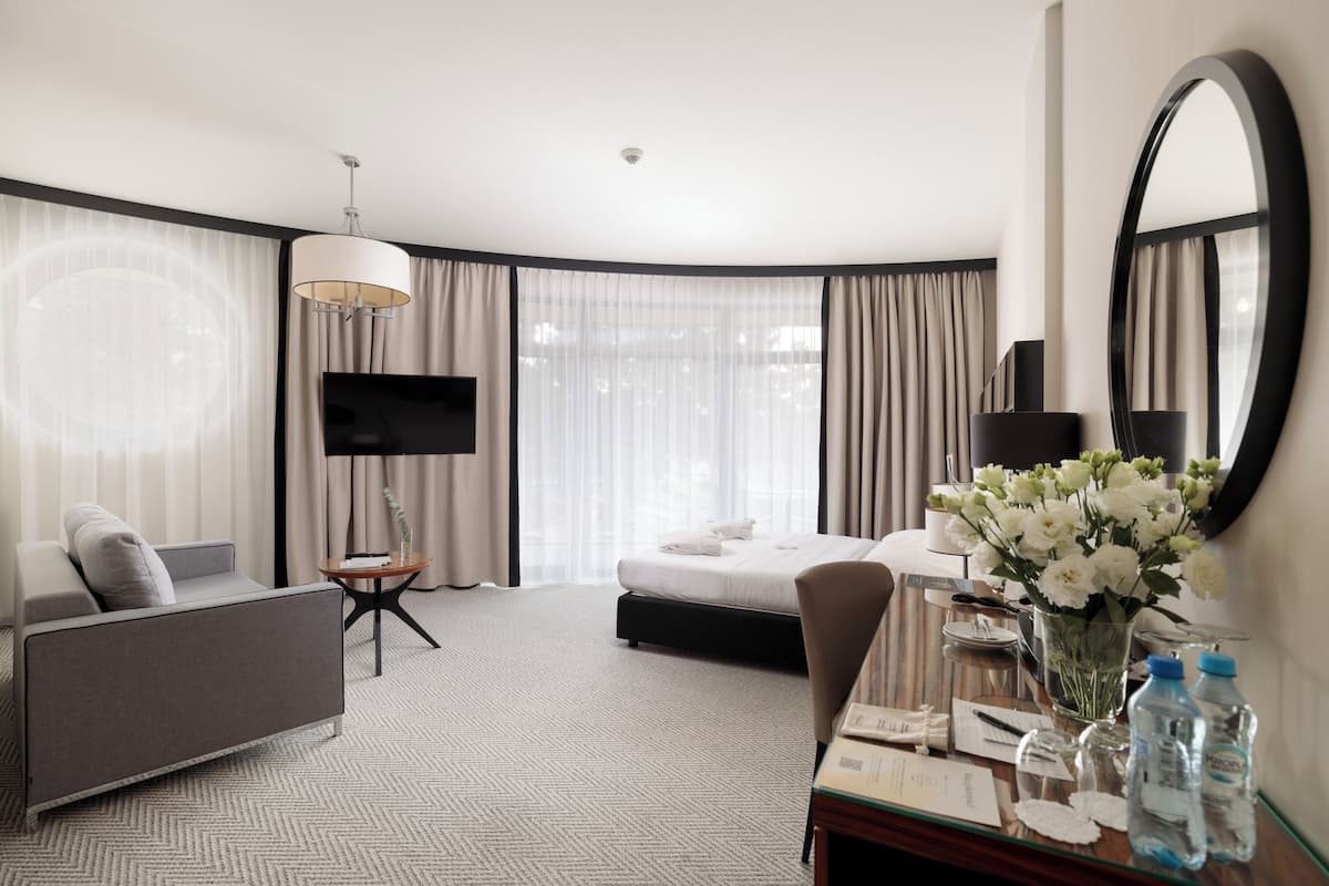 A hotel room with a bed, a sofa, a TV, a desk with a mirror, and a window with curtains.