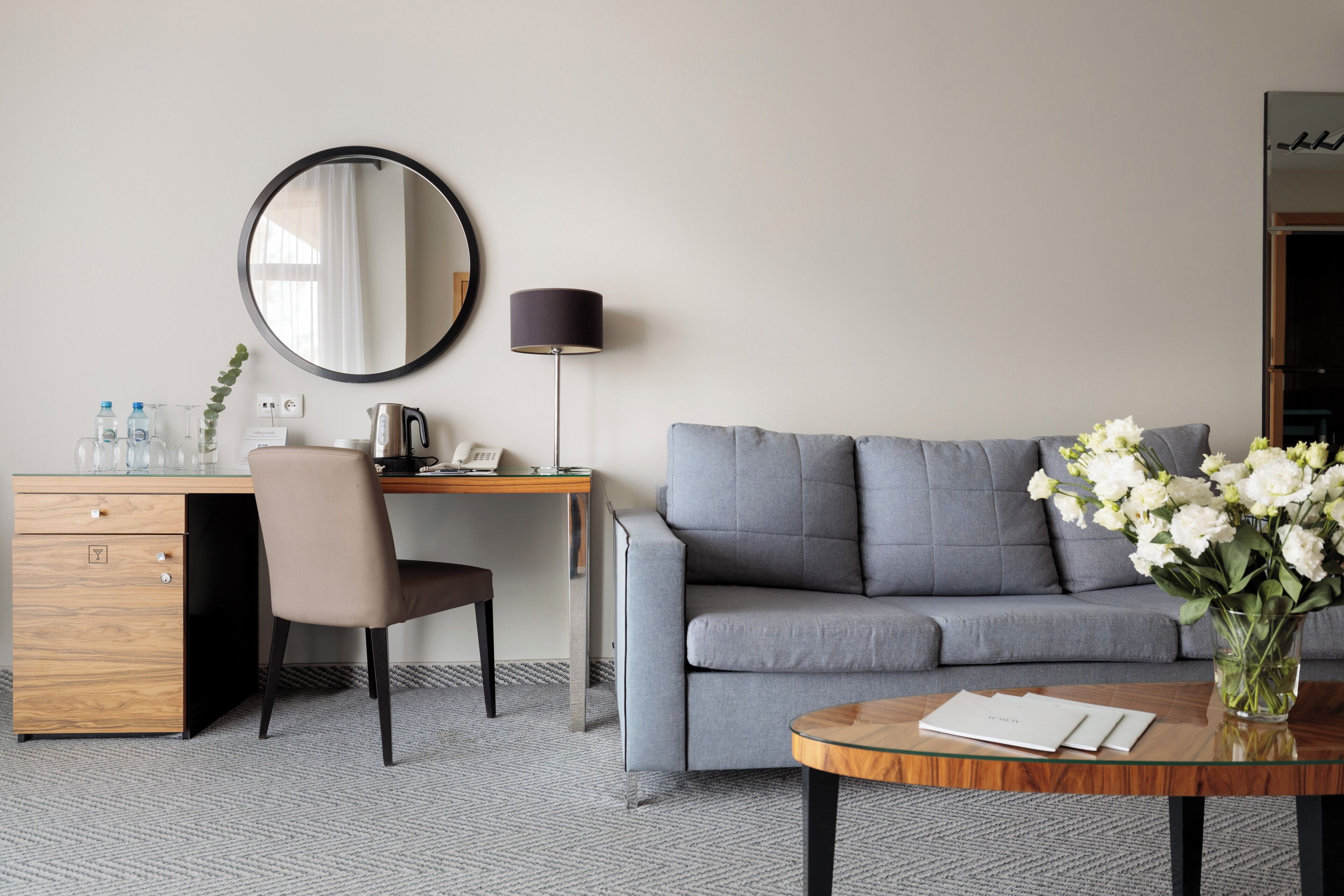A modern hotel room with a grey sofa, a wooden desk, a round mirror, a lamp, a vase with flowers, and a glass table.