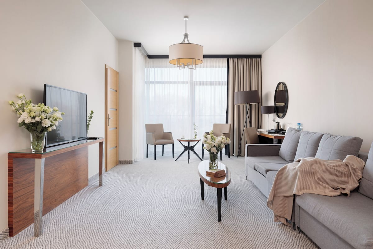 A hotel room with a TV, sofa, coffee table, and a desk with a lamp.