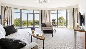 A modern living room with a sofa, armchair, and ottoman, a glass coffee table, and a large window offering a view of greenery. - Diune Hotel (Kolobrzeg)