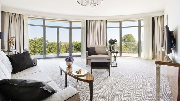 A modern living room with a sofa, armchair, and ottoman, a glass coffee table, and a large window offering a view of greenery. - Diune Hotel (Kolobrzeg)