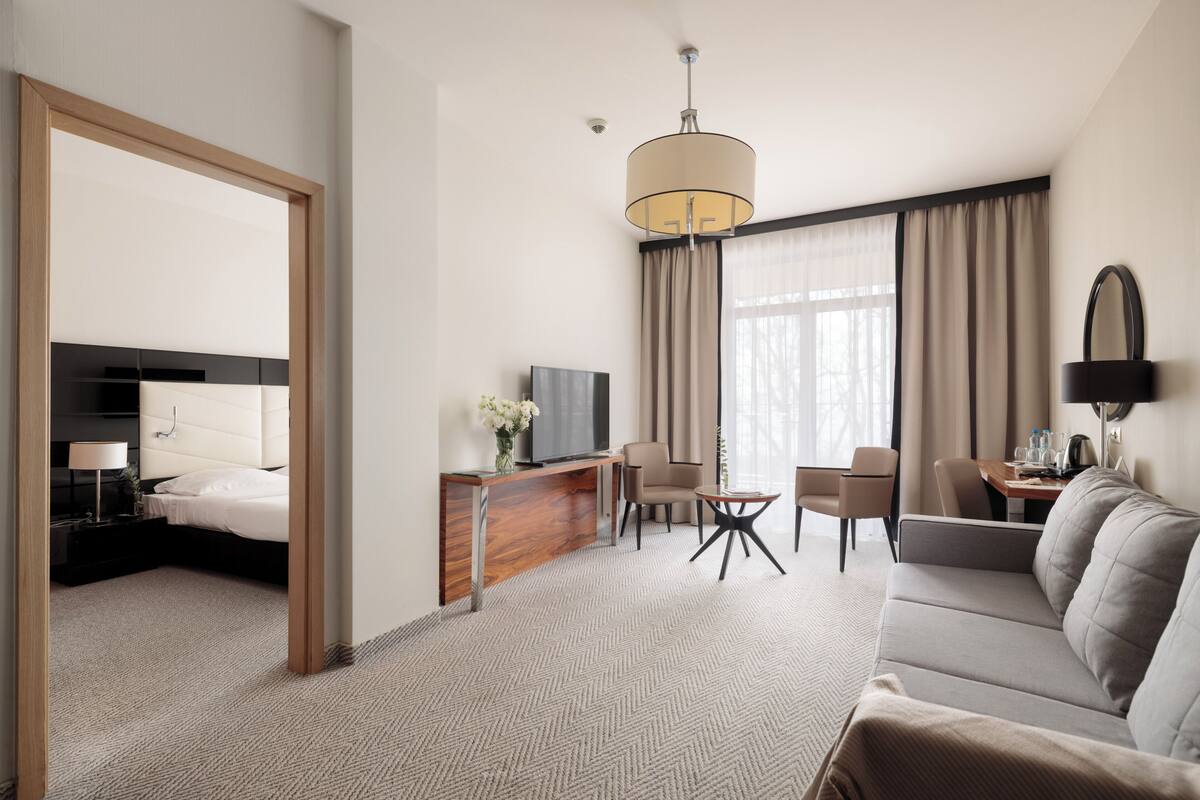 A hotel room with a bed, sofa, TV, desk, and a large window with curtains.