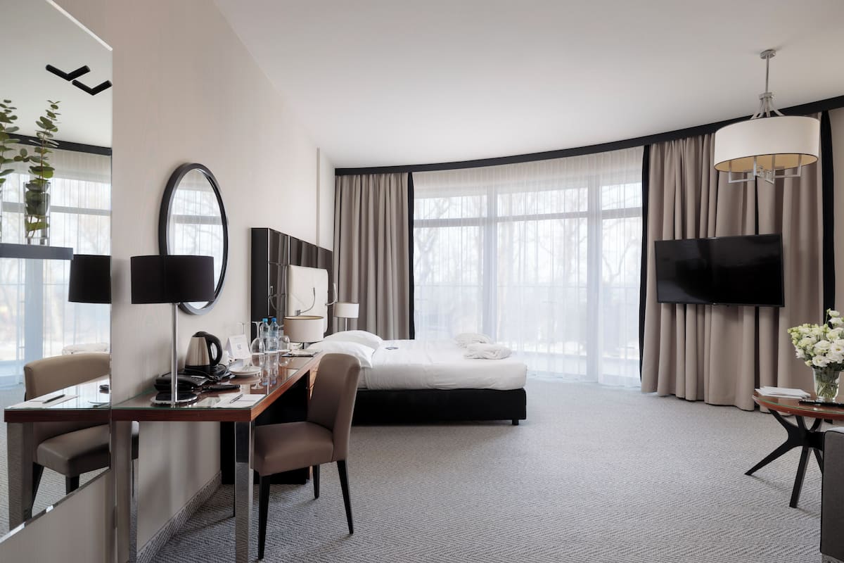 A hotel room with a bed, desk, chairs, a TV, and a large window with curtains.