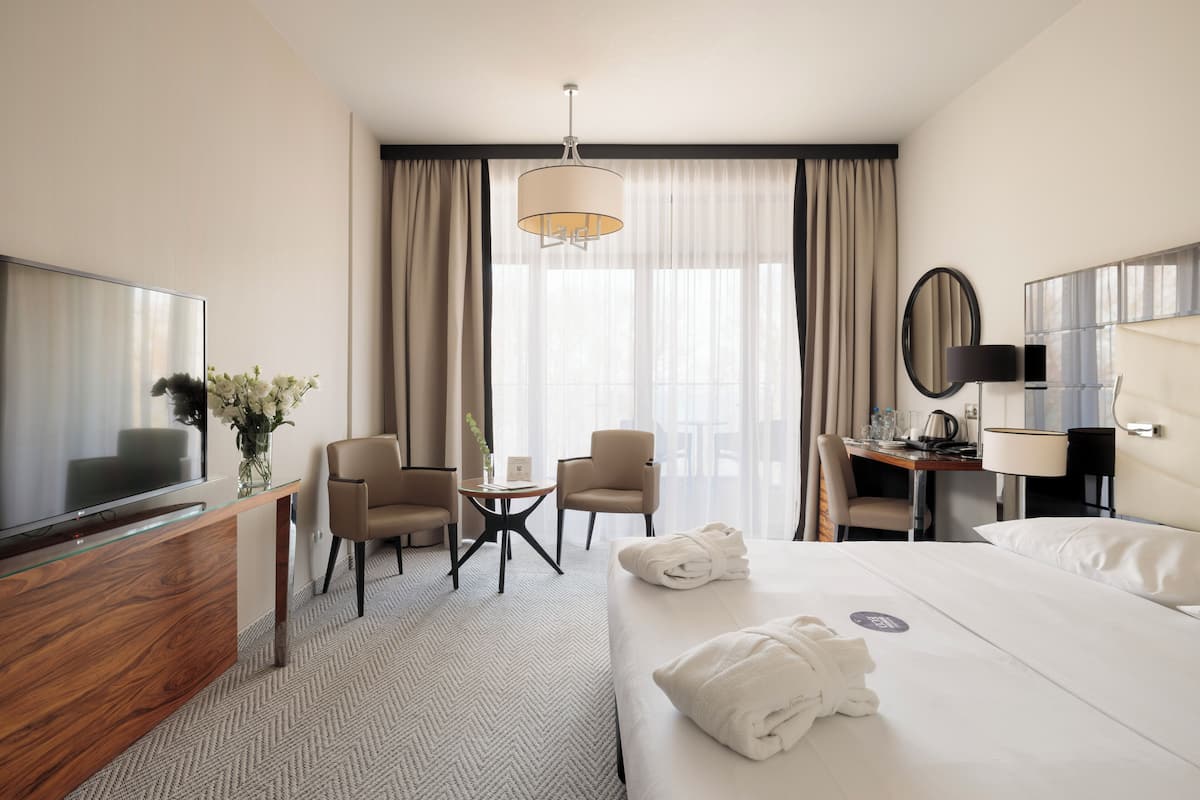 A hotel room with a bed, a desk, a TV, a chair, a lamp, and a large window with curtains.