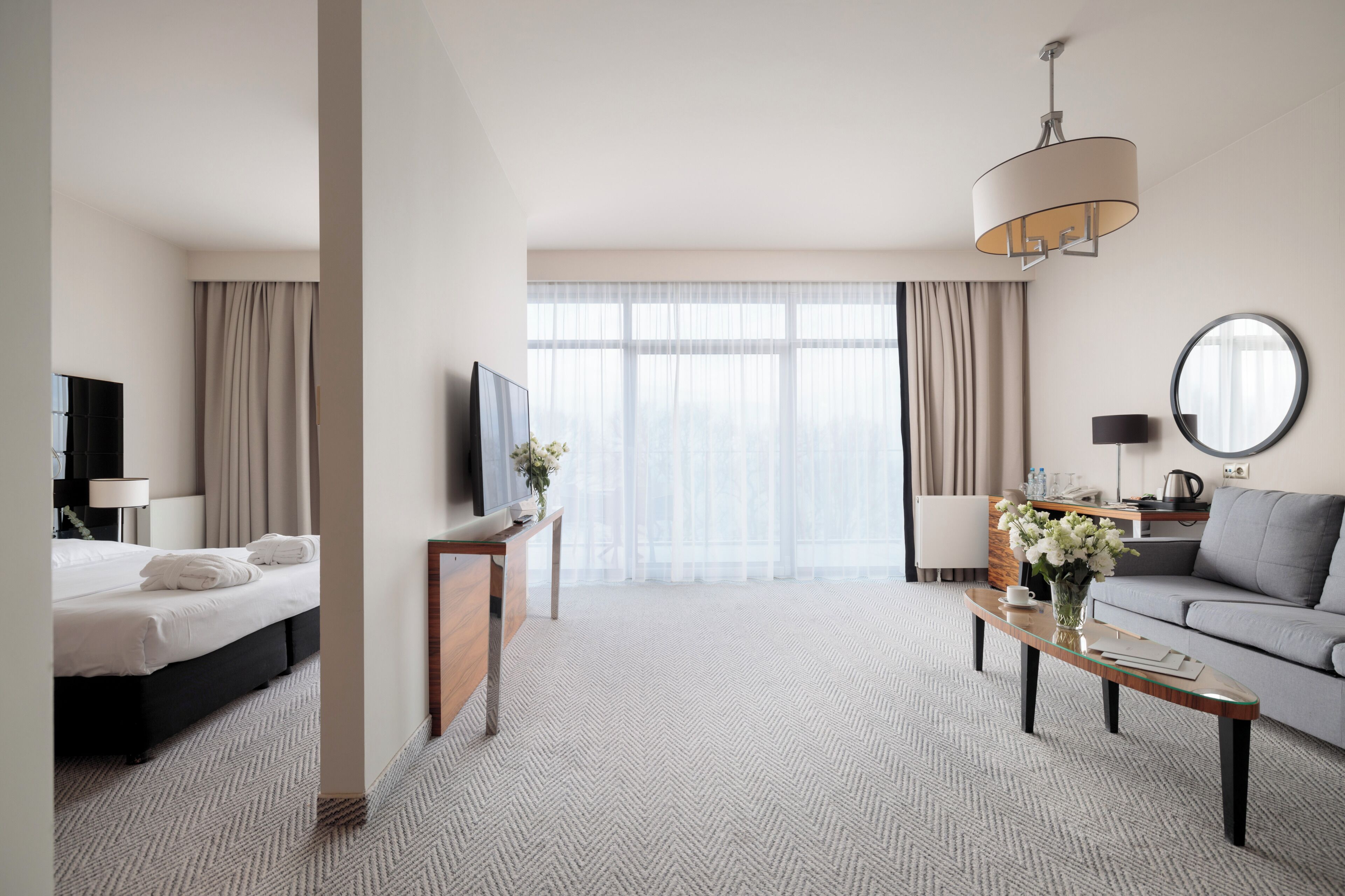 Una habitación de hotel con cama, televisión, sofá, mesa de centro y un amplio ventanal con cortinas.