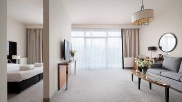 Una habitación de hotel con cama, televisión, sofá, mesa de centro y un amplio ventanal con cortinas.