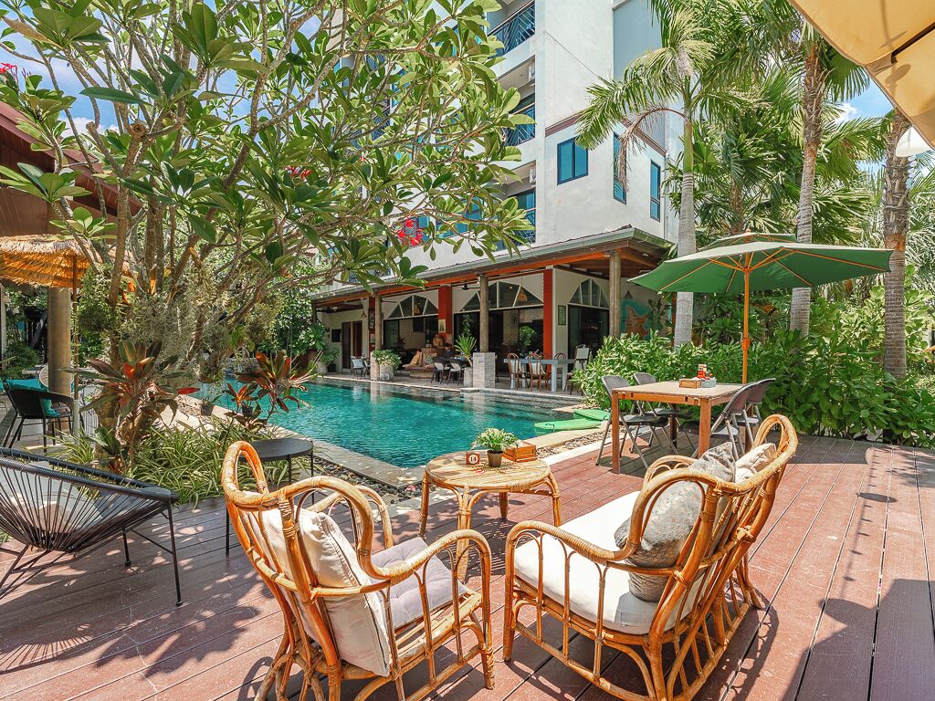 A poolside area with wicker furniture, a table with a plant, and a building with balconies in the background.
