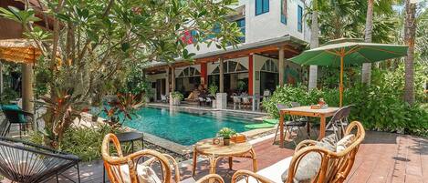 A poolside area with wicker furniture, a table with a plant, and a building with balconies in the background.