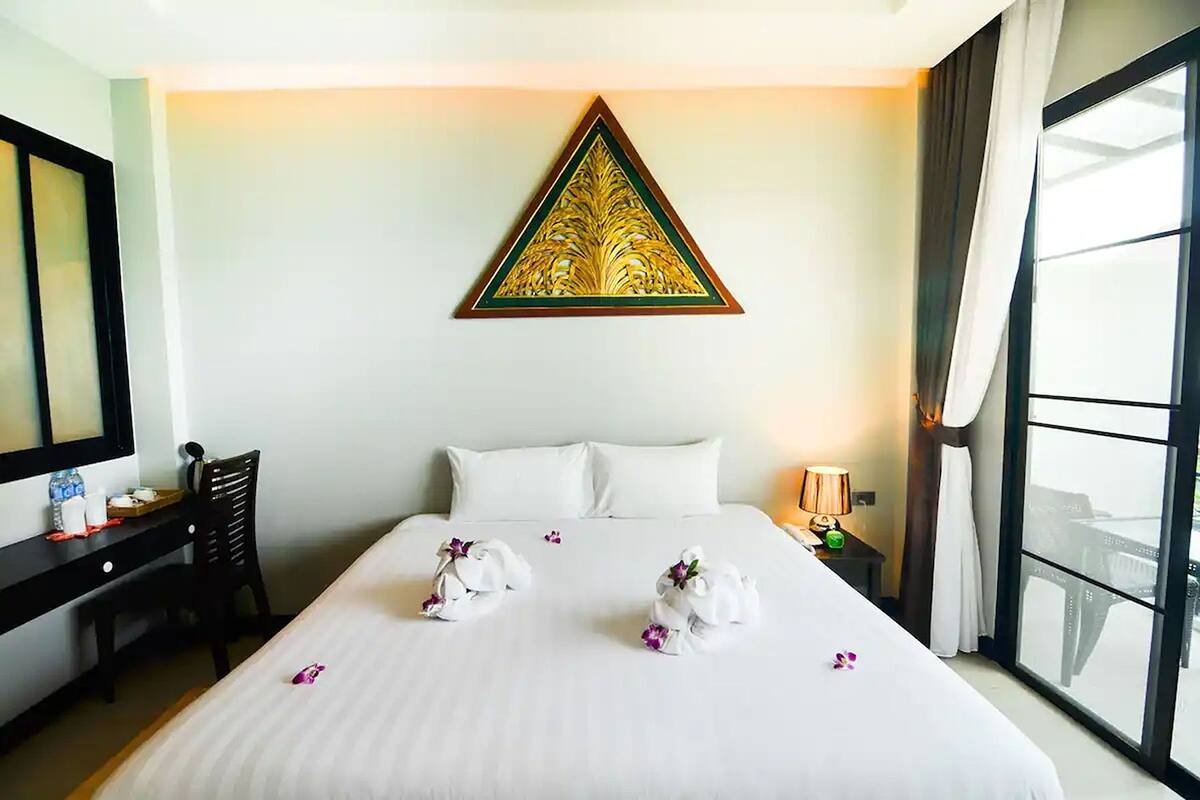 A hotel room with a neatly made bed, a desk with a chair, a large window, and a decorative wall piece.
