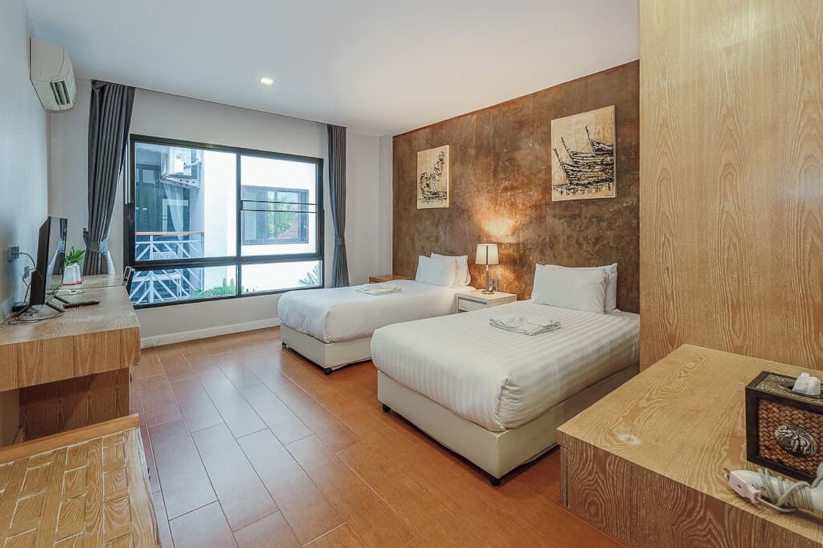 A hotel room with two beds, a desk, a chair, a window with a view of buildings, and artwork on the wall.