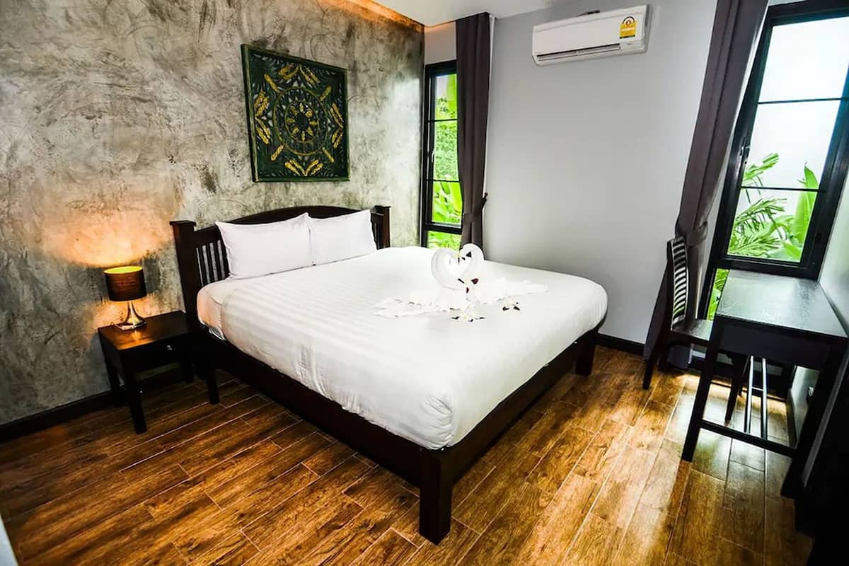 A bedroom with a wooden floor, a bed with white bedding, a nightstand with a lamp, a desk with a chair, and a window with a view of greenery.