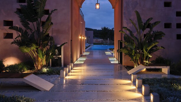 Un sendero conduce a un edificio con una entrada arqueada, rodeado de plantas tropicales e iluminado por luces colgantes.