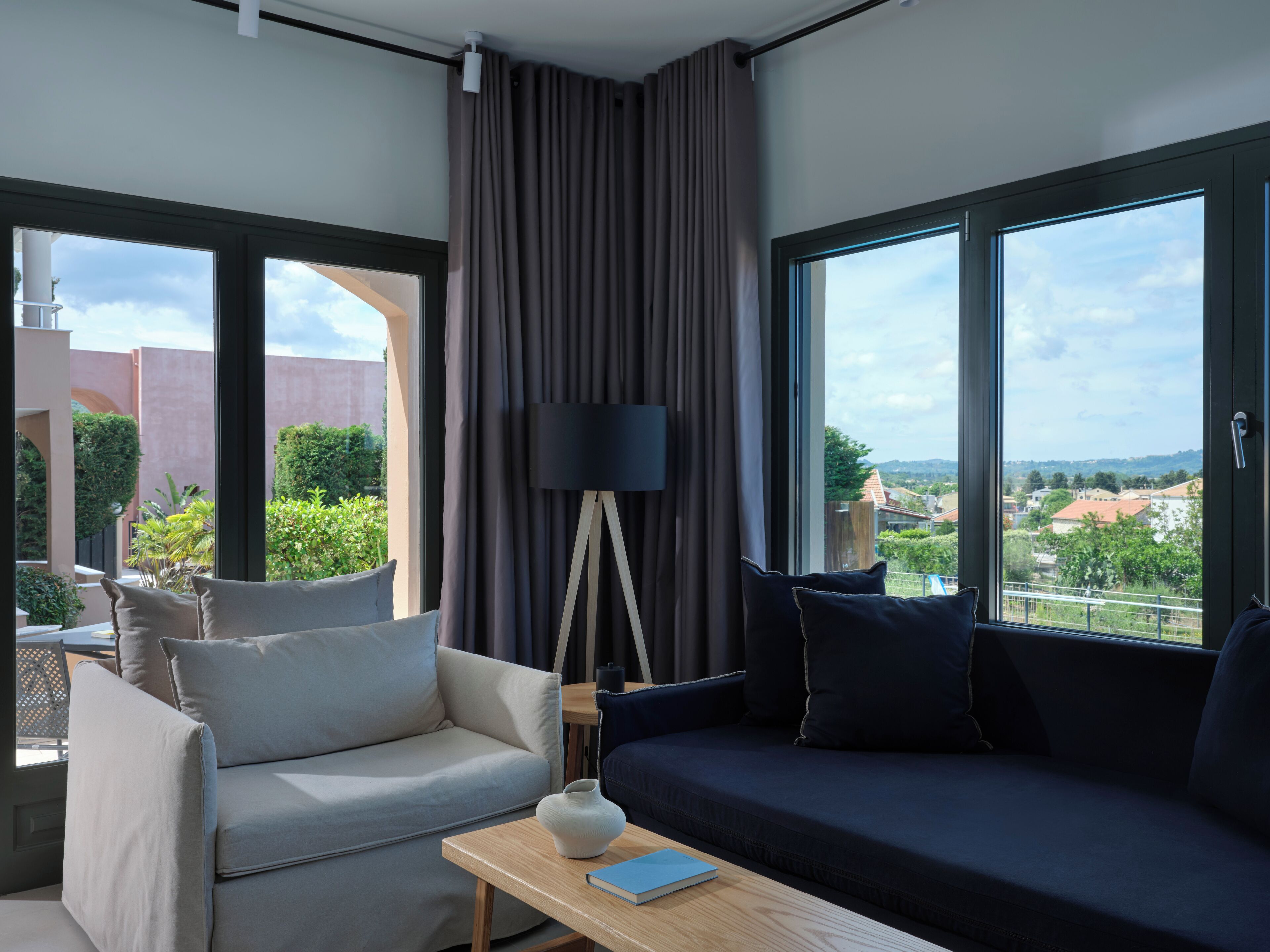 Une chambre d’hôtel moderne, dotée d’un canapé, d’un fauteuil et d’une table agrémentée d’un vase et d’un livre. De grandes fenêtres offrent une vue sur le paysage urbain.