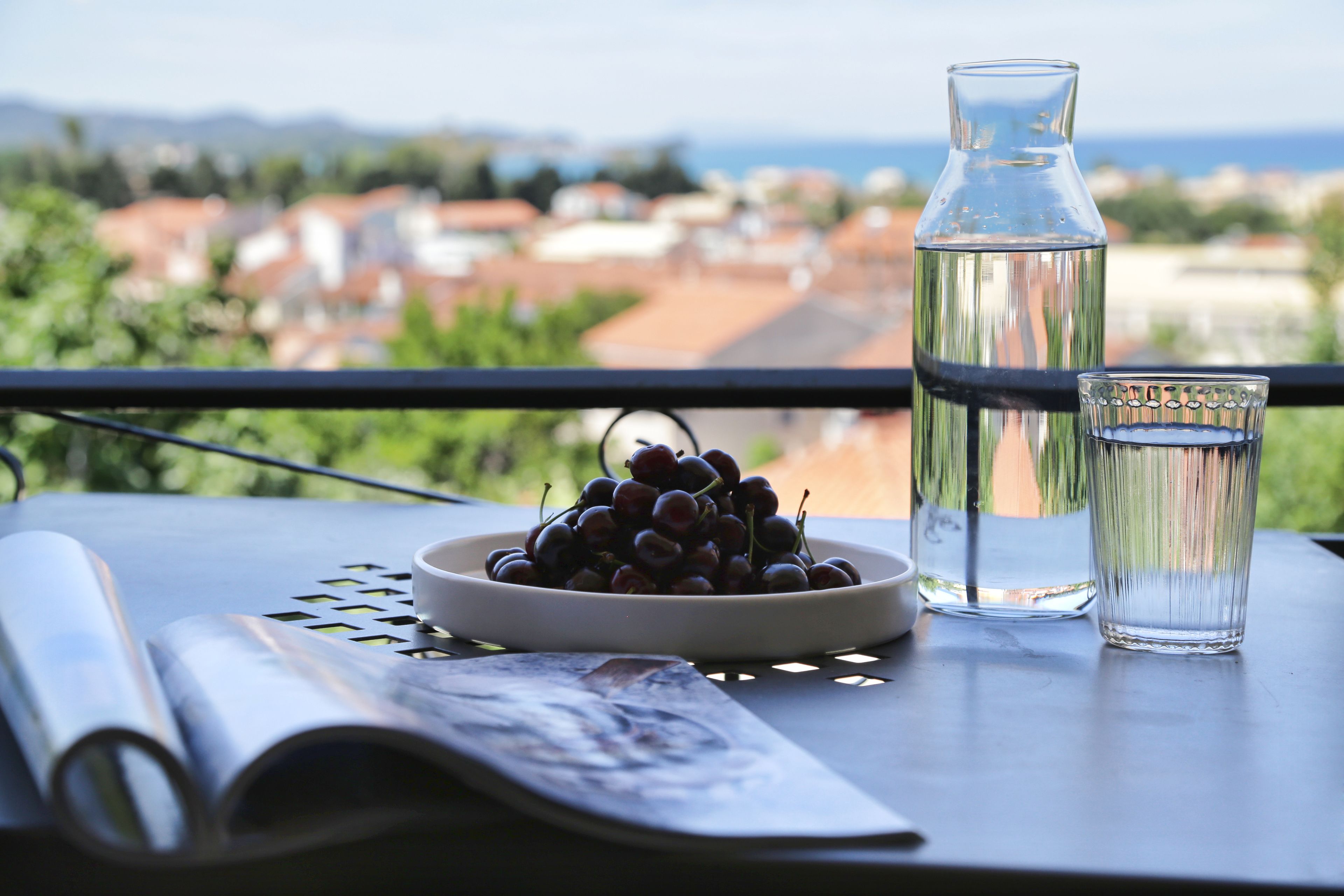 A table with a bowl of grapes, a glass of water, and a water bottle on a balcony overlooking a cityscape.