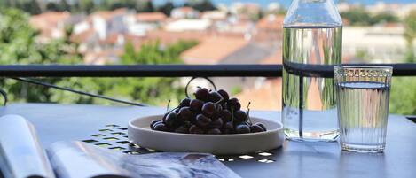 A table with a bowl of grapes, a glass of water, and a water bottle on a balcony overlooking a cityscape.
