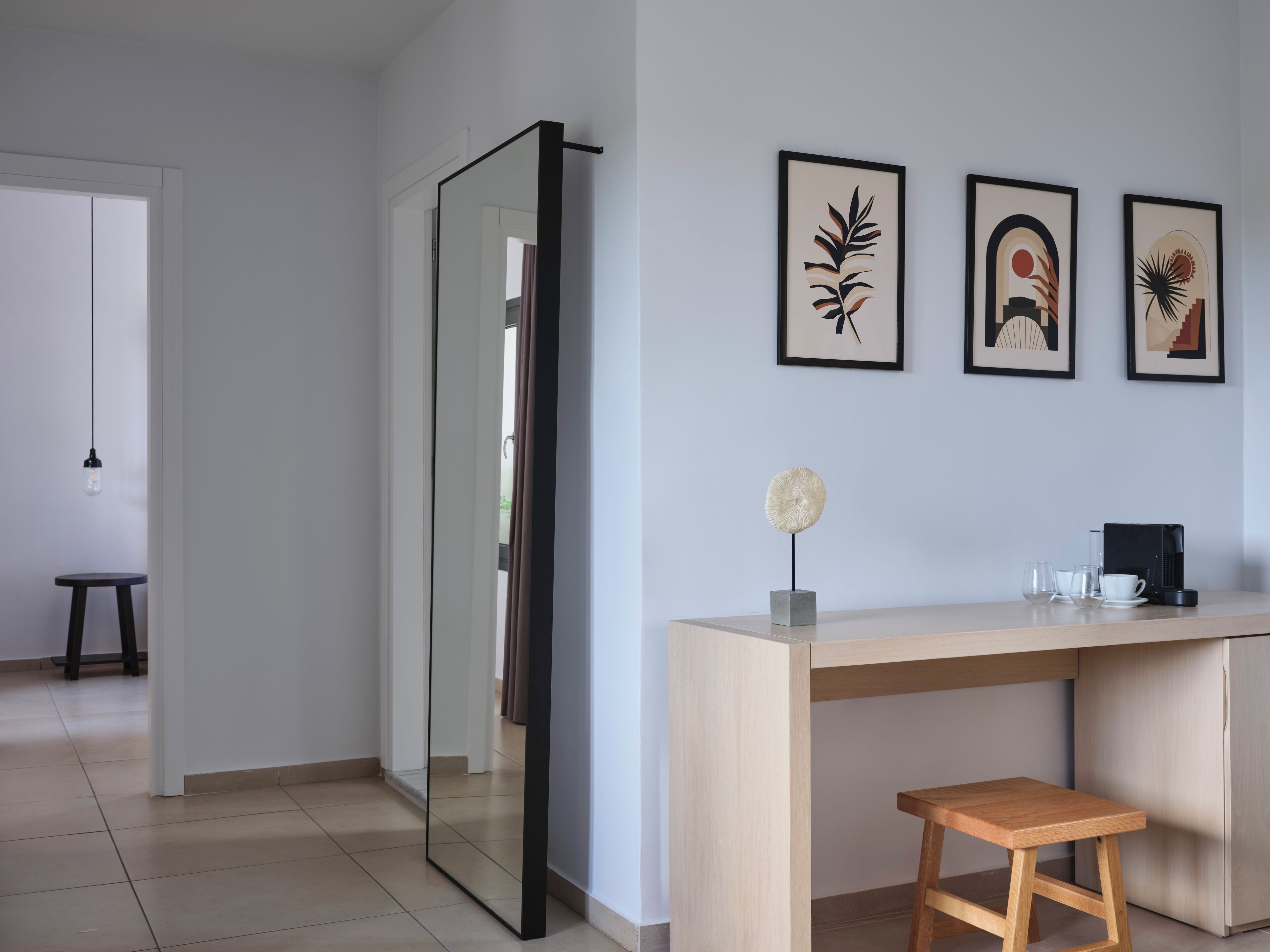 A modern kitchen with a light wood countertop, a small stool, and a coffee machine. There are framed artworks on the wall and a hanging light fixture.