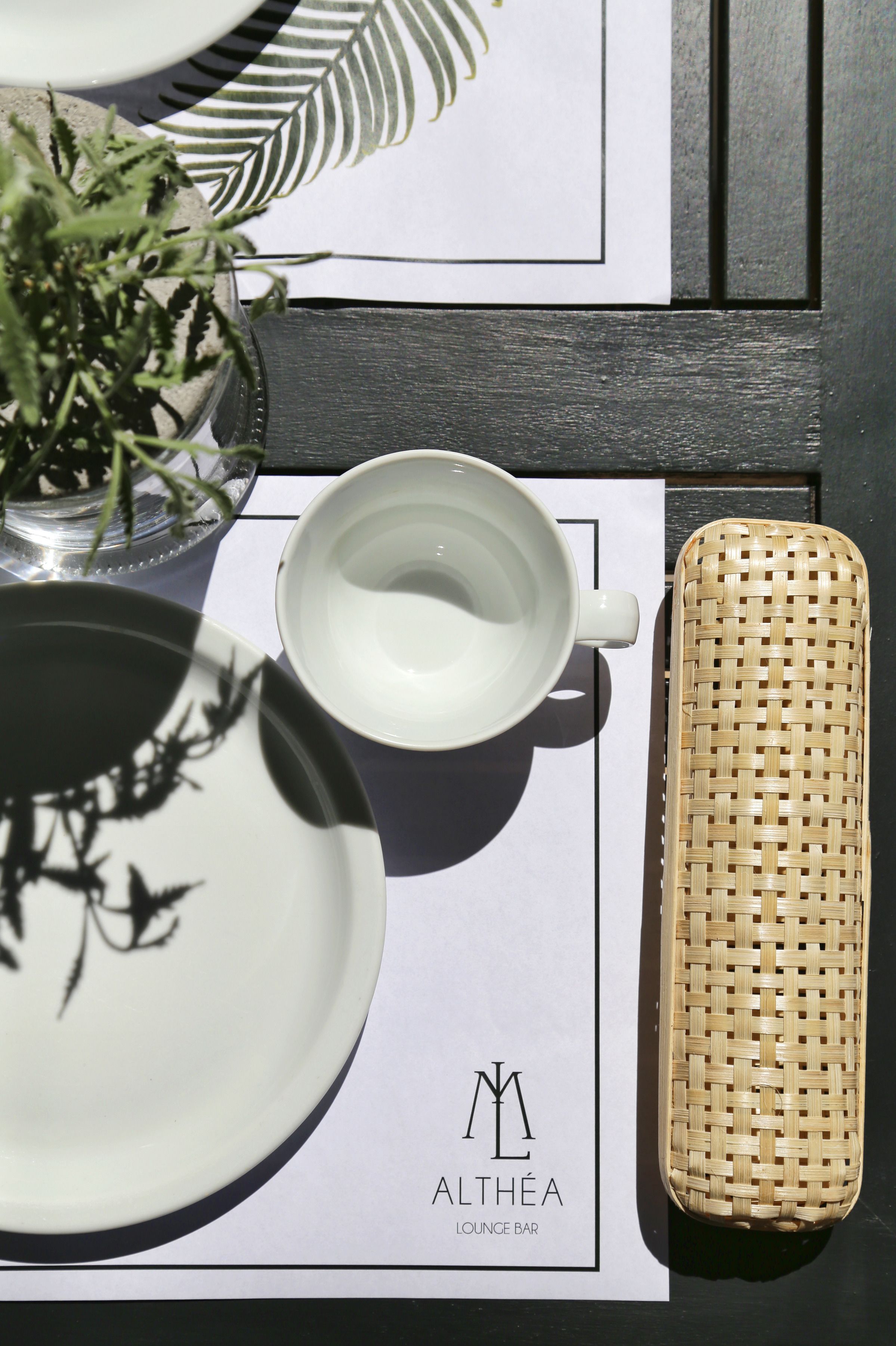 A table setting with a white plate, a white cup, a woven basket, and a potted plant. The table is on a white placemat with the logo 'ALTHEA LOUNGE BAR'.