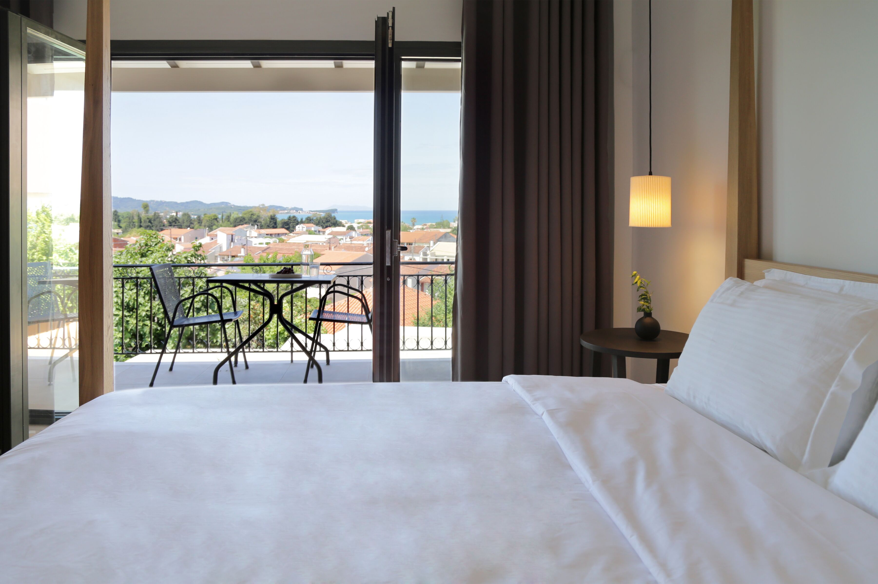 A hotel room with a bed, a small table, a chair, a potted plant, and a view of the city through a balcony.