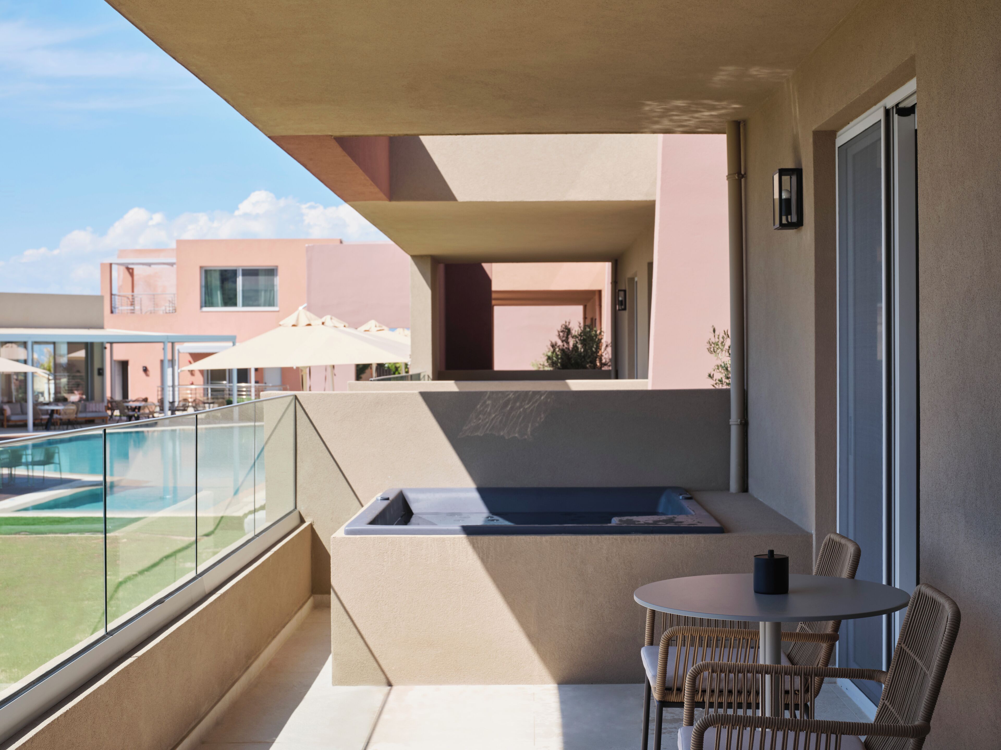 A balcony with a glass railing, a hot tub, wicker chairs, and a view of a pool and buildings.