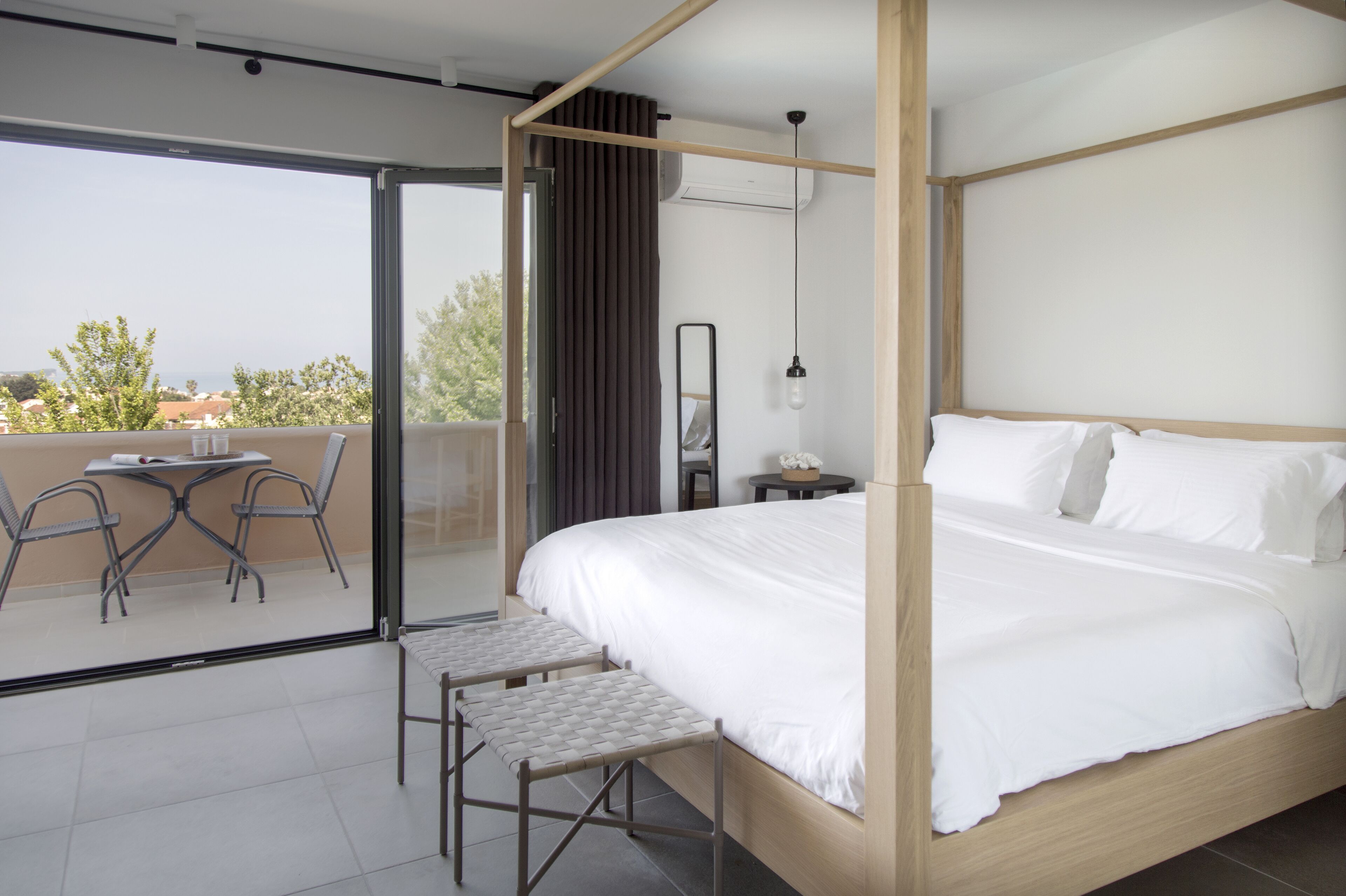 Une chambre moderne avec un lit à baldaquin, un balcon doté d’une table et de chaises, et une vue sur le paysage urbain.
