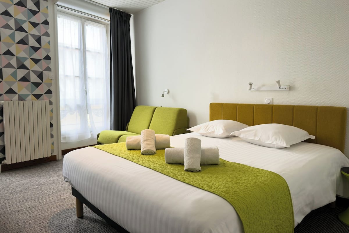 A hotel room with a large bed, a green armchair, and a window with curtains.