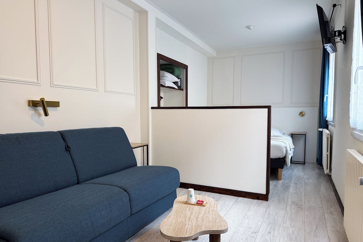 A room with a blue sofa, a small wooden table, a bed, a wardrobe, and a TV mounted on the wall.