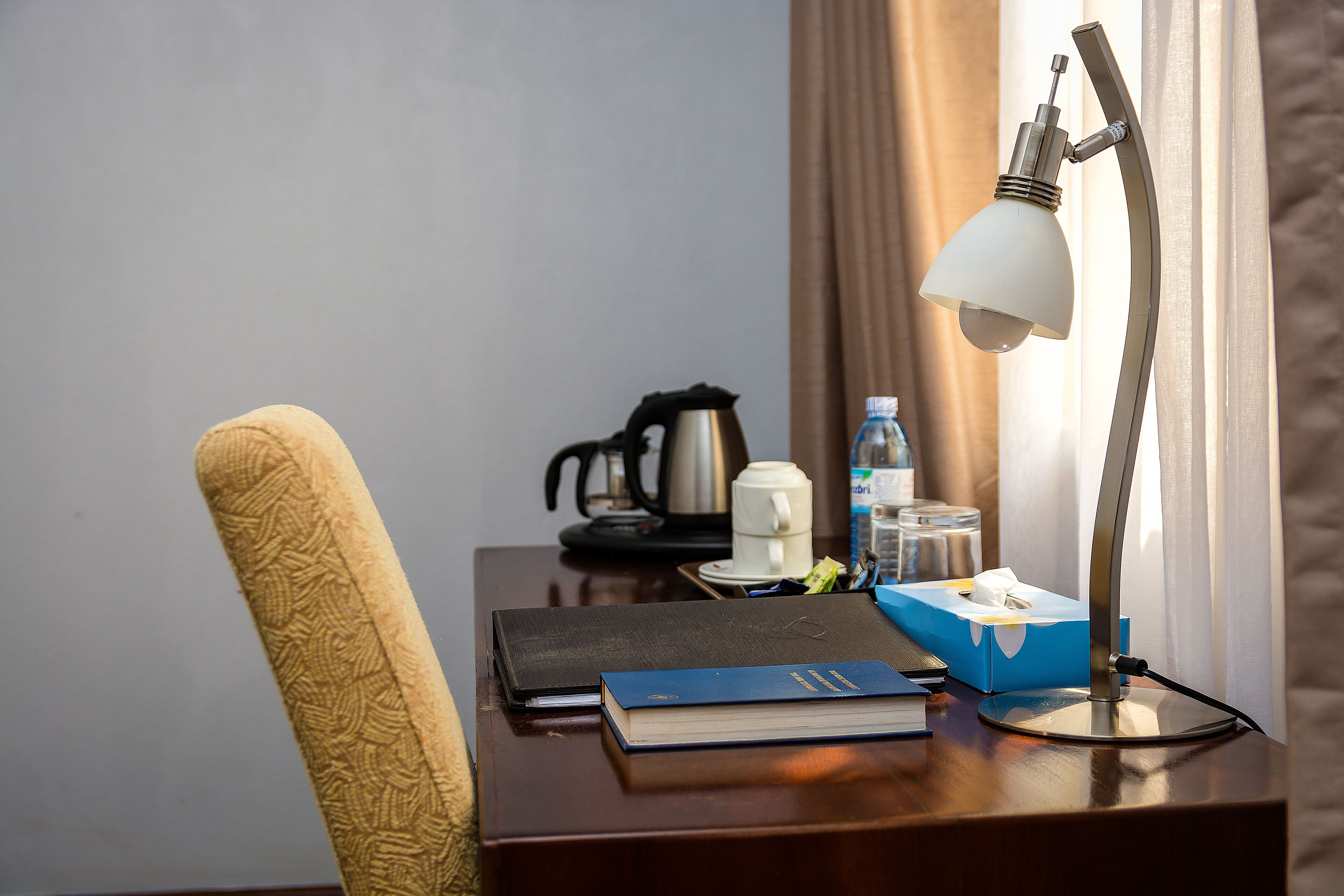 Een houten bureau met een lamp, een boek, een pak tissues, een waterfles en een koffiezetapparaat.