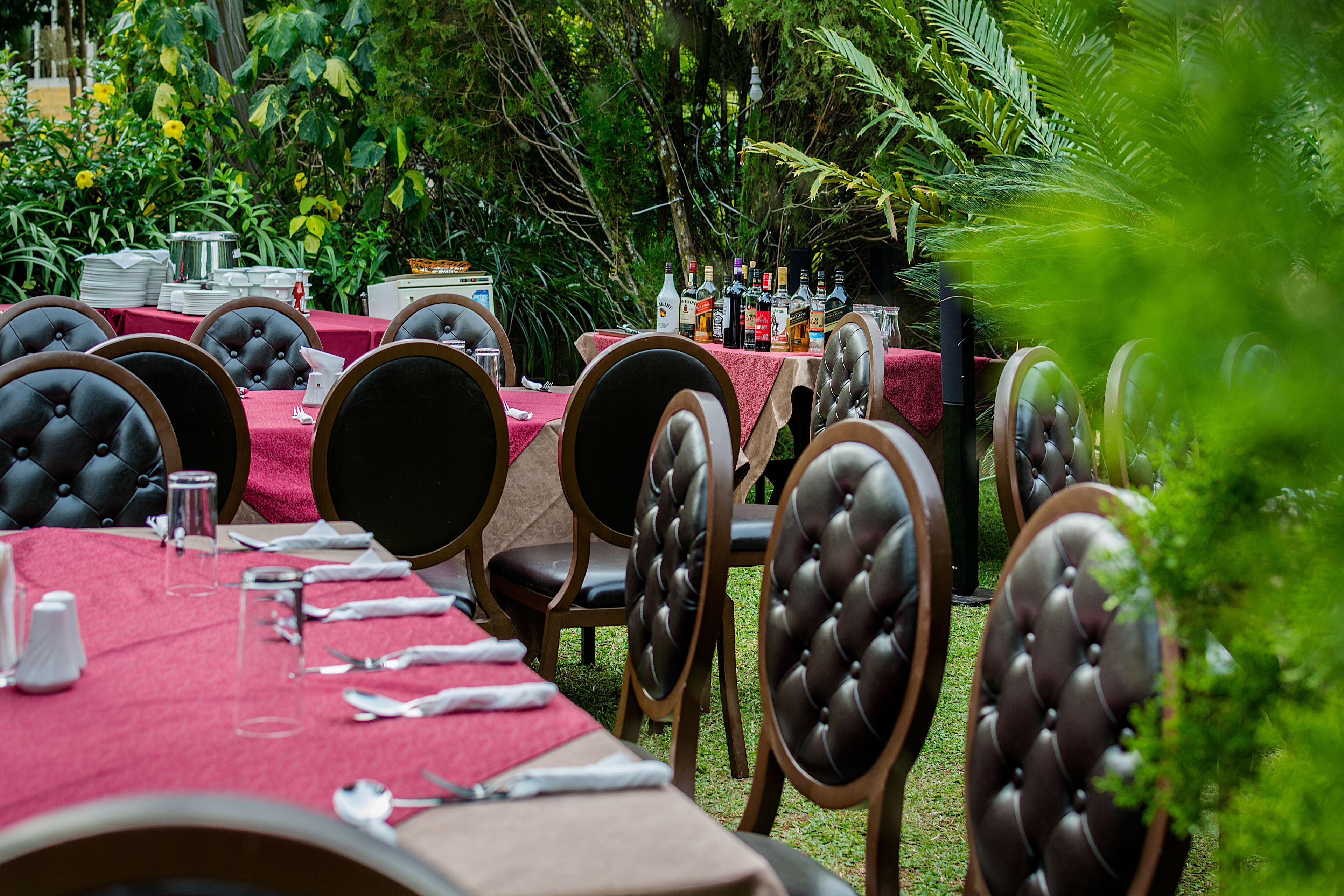 An outdoor dining area with multiple tables set with red tablecloths, black cushioned chairs, and various dining utensils.