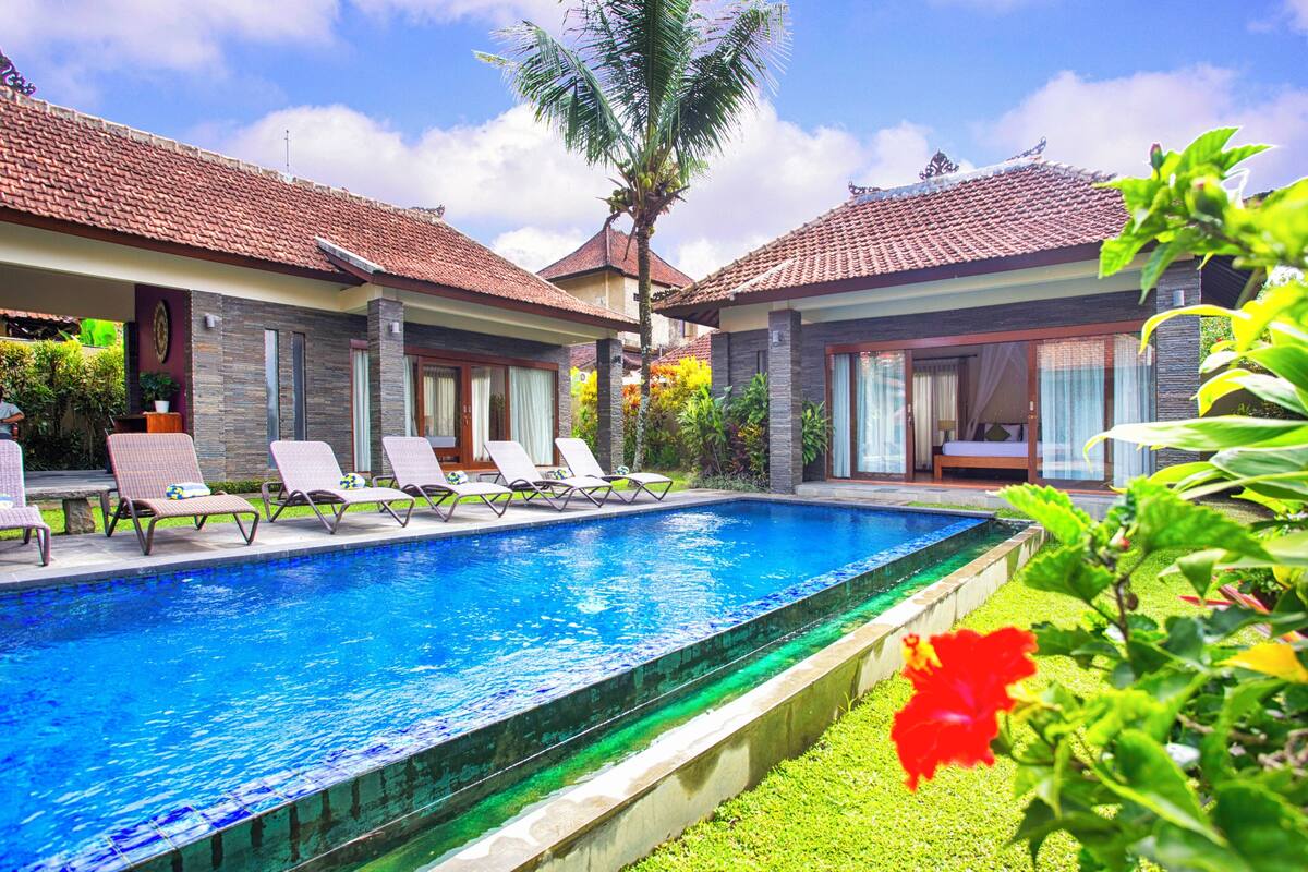A poolside view of a resort with a swimming pool, lounge chairs, and a tropical garden.