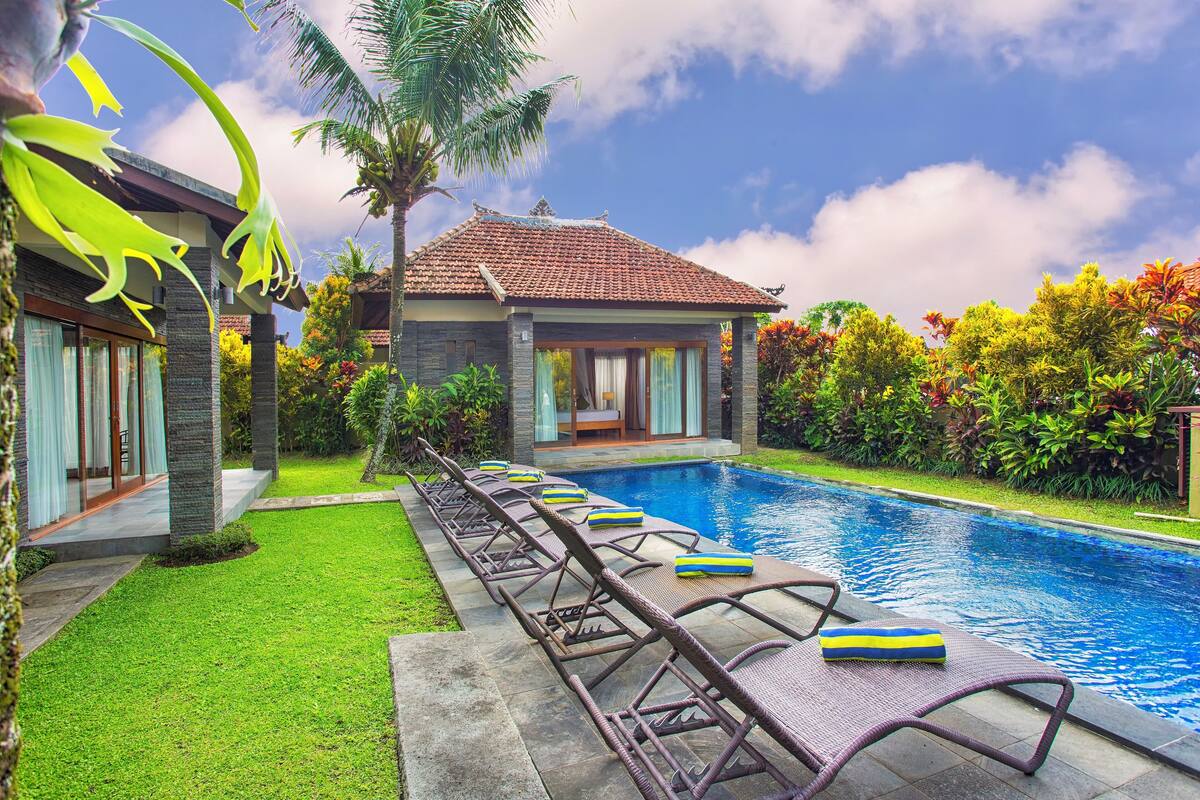 Zona junto a la piscina con sillones de descanso y una pileta, rodeada de abundante vegetación y una casa con techo de tejas.