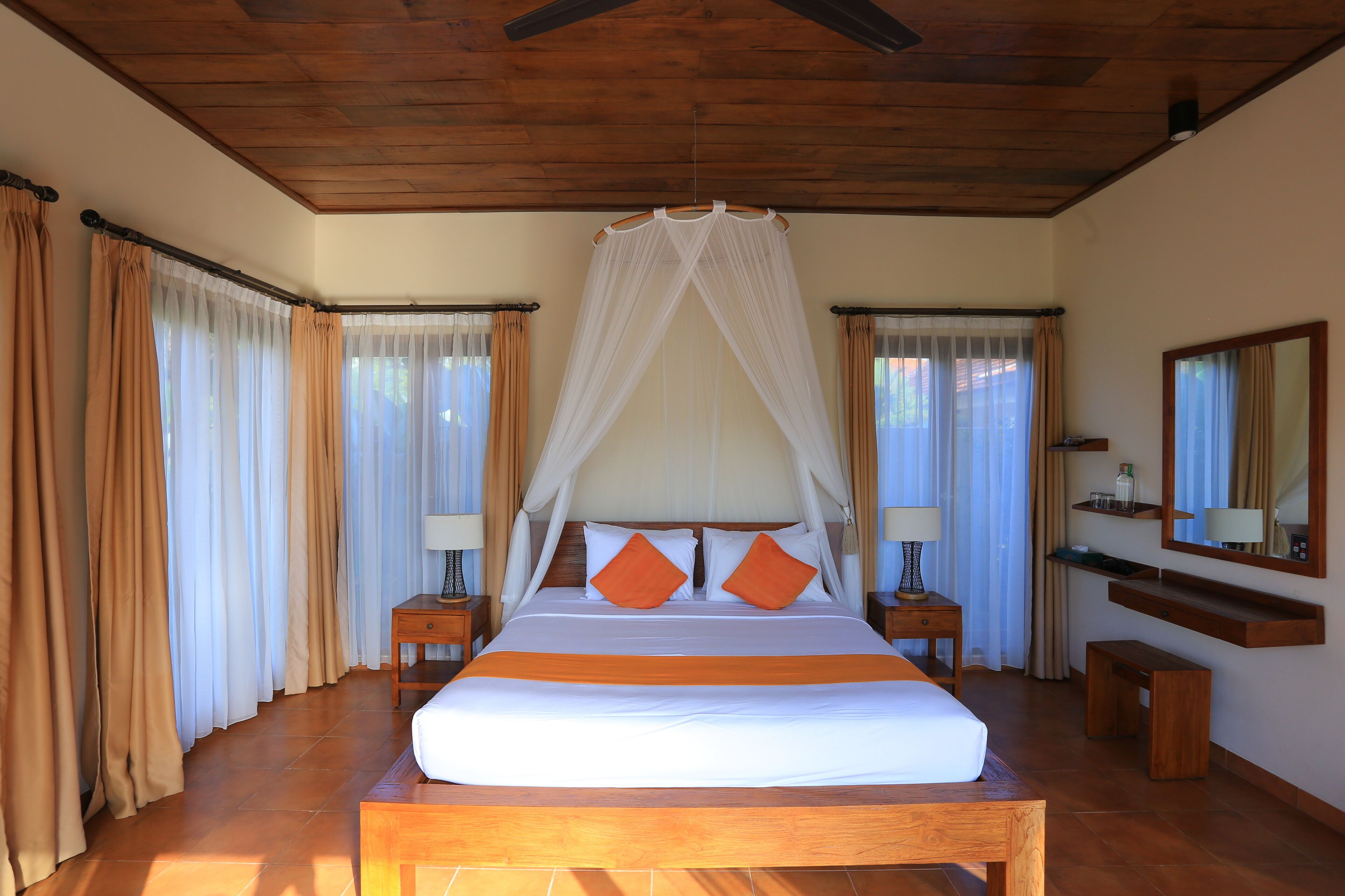 A spacious bedroom with a large bed, wooden headboard, and a canopy. There are two bedside tables with lamps, a bench, and a mirror on the wall.
