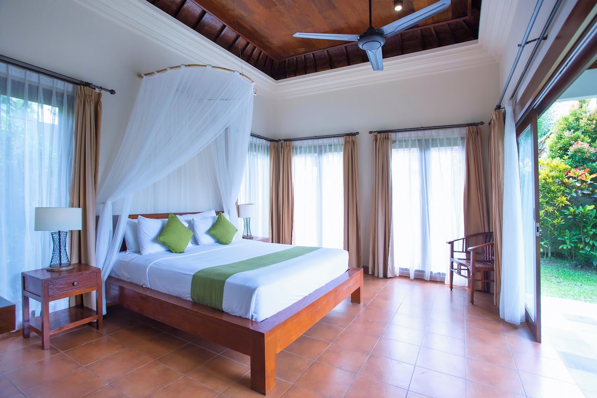 A spacious bedroom with a large bed, a ceiling fan, and a view of the garden through the windows.