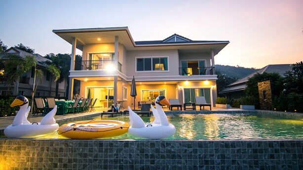 Una casa moderna de dos pisos con piscina, flotadores con forma de cisne y un área de comedor con mobiliario exterior.